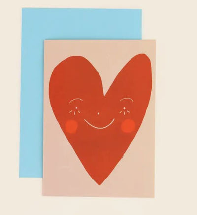 A Wonky Heart Greeting Card featuring a large red heart with a smiling face and rosy cheeks on a beige background. The card, from the Blue Dog Posters collection, sits atop a light blue envelope.