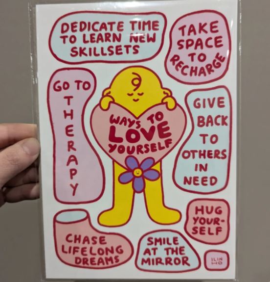 A hand holds the "Ways to Love Yourself" A5 Archival Print by Inner Baby from Blue Dog Posters, featuring a yellow figure with a heart and self-care tips in vivid red and pink bubbles—perfect for nurturing your inner child.
