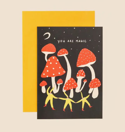 The You are Magic Greeting Card by Blue Dog Posters showcases a black background adorned with red and white mushrooms, along with four yellow figurines holding hands and dancing. A crescent moon and stars glimmer in the Manchester, UK sky above. The words "You are magic" embellish the top, complemented by a yellow envelope included behind it.