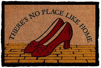 Part of our Homewares Collection, the MTV Wizard of Oz No Place Like Home coir door mat features the text "THERE'S NO PLACE LIKE HOME" alongside an illustration of red high-heeled shoes on a yellow brick road background, adding a whimsical touch to your entryway.