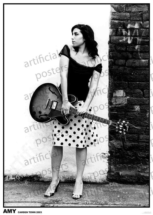 Black and white photo of a woman standing against a white wall with a brick section. She holds a guitar and is dressed in a polka-dot skirt and dark top. The image, printed on premium gloss paper, reads "AMY" and "CAMDEN TOWN 2003." Perfect for any music collection poster. Titled *Amy Winehouse - Camden*, this piece is brought to you by MUS 291.