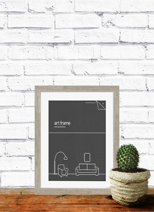 A framed minimalist poster featuring a couch and a lamp against a charcoal background is placed on a wooden surface. Highlighted in Blue Dog Posters' Deluxe Frame A3 Ash (double mat board), it sits beside a small potted cactus. The backdrop is a white painted brick wall.