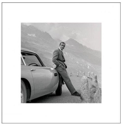 Black-and-white image of a person standing next to an old-fashioned car on a mountain road. The individual is wearing a suit and leaning against a rock barrier, with mountains visible in the background. Part of the MEZ 248 Contemporary Art Collection, this piece captures timeless elegance in nature with its James Bond - Aston Martin Square.