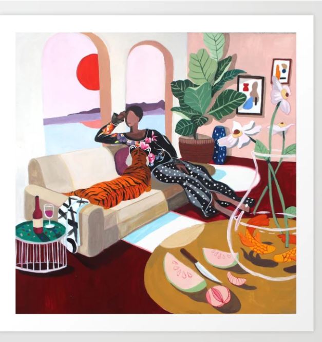 In a vibrant room, someone relaxes on a sofa with the Tiger Queen by Artyguava from GAL19, surrounded by tropical decor and houseplants. They enjoy a drink near an Art Print, with a wine bottle, glass, and fruit bowl on the side table. A large window offers views of a sunset over water.