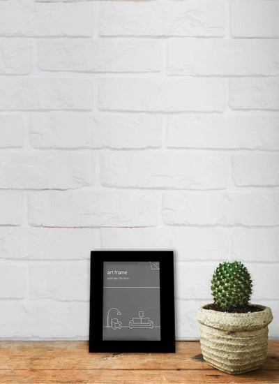 A small black frame, the Classic Frame 10 x 15cm Black by Blue Dog Posters, sits on a wooden surface against a white brick wall. Next to it, there is a small potted cactus in a woven pot. The setting has a simple, modern, and cozy feel, emphasizing the charm of classic frames in contemporary decor.