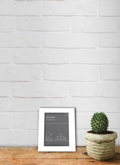 A white Classic Frame 10 x 15cm White by Blue Dog Posters with a minimalist design rests on a wooden surface against a white brick wall. Next to the Classic Frame 10 x 15cm White is a potted cactus in a textured, beige pot. The scene has a clean, modern aesthetic.
