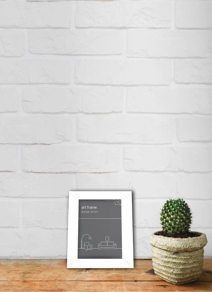 A white Classic Frame 10 x 15cm White by Blue Dog Posters with a minimalist design rests on a wooden surface against a white brick wall. Next to the Classic Frame 10 x 15cm White is a potted cactus in a textured, beige pot. The scene has a clean, modern aesthetic.