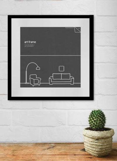 A framed picture from the Blue Dog Posters Deluxe Frame 40x40cm Black hangs on a white brick wall. The picture, set in a black frame, shows a simple line drawing of a living room with a lamp, armchair, and sofa. Below the picture, a small cactus in a textured pot sits on a wooden surface.