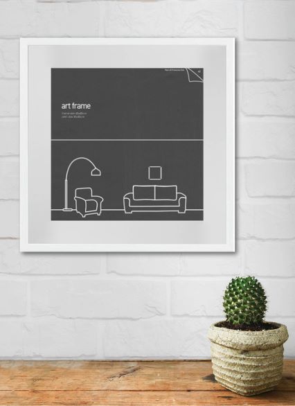 A framed minimalist black-and-white art piece from Blue Dog Posters' Deluxe Frame Collection, measuring 40x40cm with a white frame, hangs on a white brick wall. The art displays a simple line drawing of a living room with a floor lamp, an armchair, and a sofa. A potted cactus sits on a wooden surface beneath the frame.