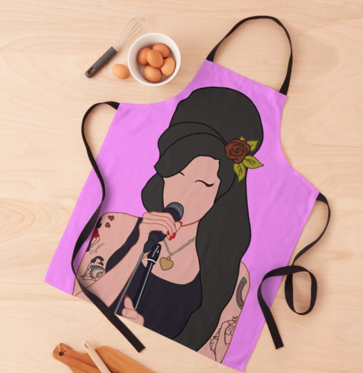 A colorful Blue Dog Posters Amy Winehouse Apron lies on a wooden surface, featuring a sublimation printed illustration of a woman with long black hair, a flower in her hair, and tattoos on her arms, singing into a microphone. Nearby are a whisk, cracked eggs in a bowl, wooden utensils, and a cutting board.