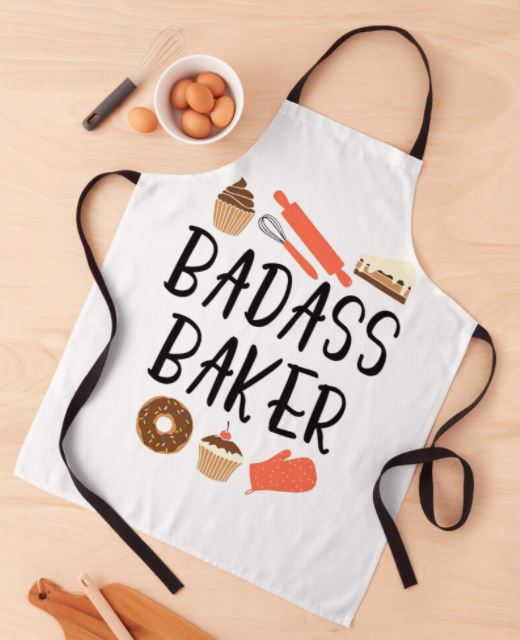A white apron with black neck and waist ties lies on a wooden surface. This stylish kitchen essential, the Badass Baker Apron from Blue Dog Posters, features the words "BADASS BAKER" in bold letters and illustrations of baking items such as a rolling pin, whisk, whisking bowl, pie, donut, cupcake, and an oven mitt.