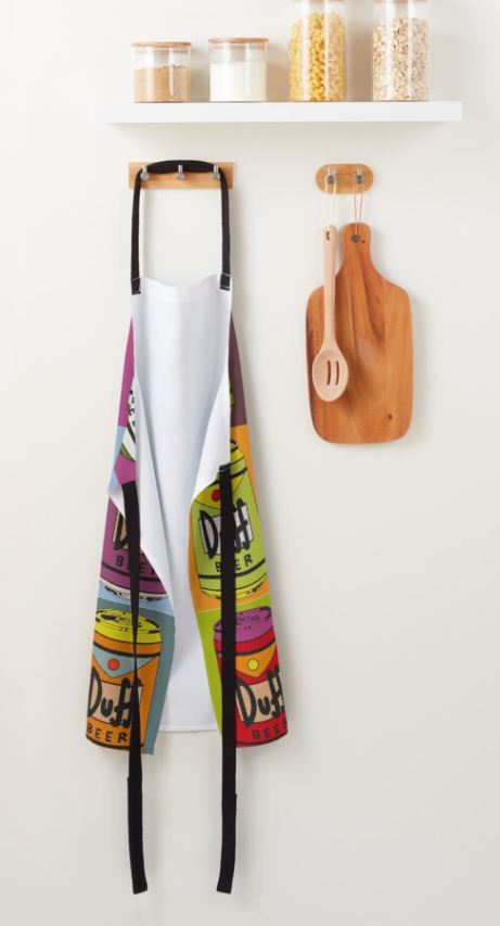 A vibrant Warhol Duff Beer Apron from Blue Dog Posters, conveniently machine washable, hangs on a hook beneath a wooden shelf. The shelf above holds three glass jars filled with various pantry items. To the right of the apron, a wall hook displays a small wooden cutting board and wooden spoon from the Homewares Collection.