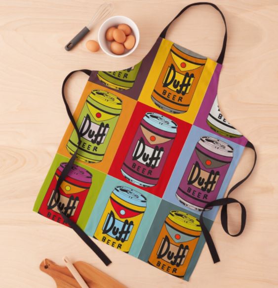 Part of the Blue Dog Posters Homewares Collection, the Warhol Duff Beer Apron features a vibrant pop art design with multiple cans of "Duff Beer" in bright colors. Set against a light wooden surface, it's accompanied by kitchen utensils like a whisk, eggs, and a wooden cutting board and is machine washable for easy care.