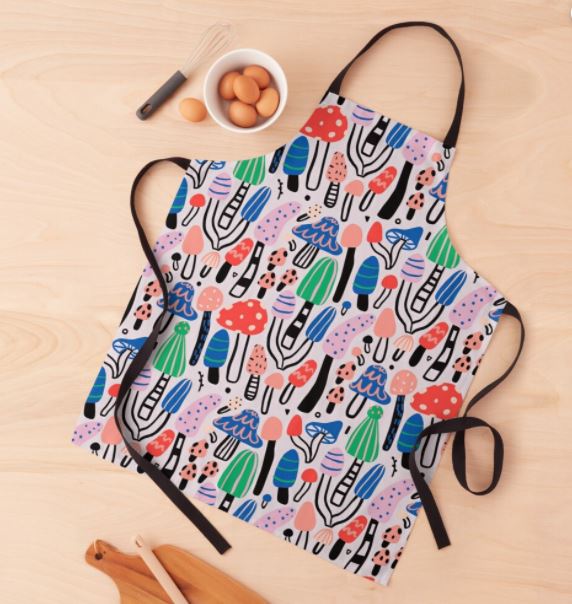 The Bold Cool Mushrooms Apron from Blue Dog Posters, featuring a whimsical pattern of various mushrooms, plants, and abstract shapes in vibrant greens, reds, blues, and pinks, lies on a wooden surface near a bowl of eggs and a whisk. Part of our Homewares Collection, the apron boasts black straps and a durable neck band.