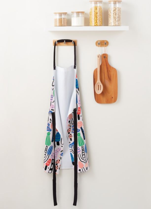 A Bold Cool Mushrooms Apron by Blue Dog Posters, featuring vibrant colors and a sturdy neck band with abstract patterns, hangs on a wall hook beside a wooden cutting board and spoon. Above, a white shelf from our Homewares Collection holds four clear jars filled with grains and pasta against a plain, light-colored wall.