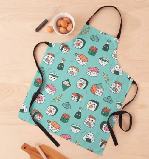 A Kawaii Japanese Sushi Apron from Blue Dog Posters, featuring a teal color and a playful sushi-themed design, is laid out on a light wooden surface. Made from 100% polyester and machine washable, this apron sits next to kitchen items including a wire whisk and a small bowl filled with eggs.