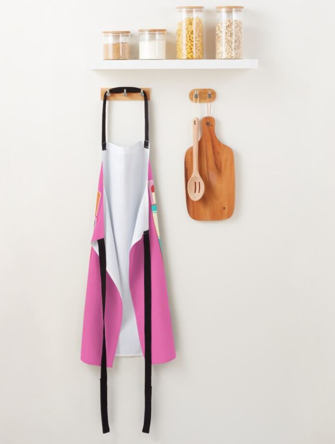 A kitchen wall with a white shelf holding jars filled with pasta, oats, and flour. Below, a vibrant "I Think You Are Hot Apron" from Blue Dog Posters with black straps hangs next to a wooden cutting board and utensils, including a spoon and fork, displayed on smaller wooden hooks. The apron is machine washable.
