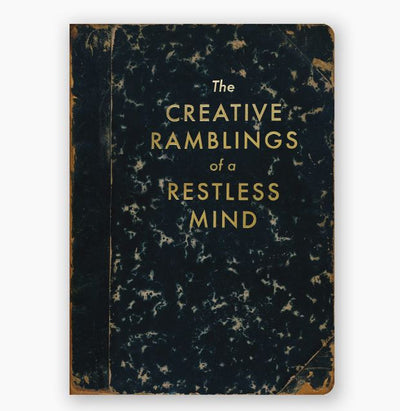 The Creative Ramblings Journal by Blue Dog Posters is a black notebook with scuffed corners, featuring the title "The Creative Ramblings of a Restless Mind" in gold foil stamped letters. Its marbled texture on the cover emphasizes its aged and well-used appearance. Despite its history, this practical journal still has numerous blank pages ready for your inspiration.
