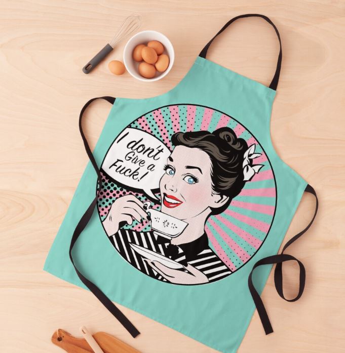 The "I Don't Give a Fuck Apron" by Blue Dog Posters is a teal, machine-washable polyester apron adorned with an illustration of a smiling, retro-style woman holding a paper that reads "I don't give a fuck!" in comic book font. The apron is displayed on a wooden surface alongside a whisk, bowl of eggs, and wooden cooking utensils.