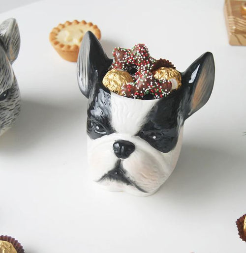 A whimsical Ceramic Planter - Frenchie from Blue Dog Posters, designed as the black-and-white head of a French Bulldog, brims with delectable chocolate treats. Surrounding the charming container are an array of small desserts, including tarts and chocolate truffles, all elegantly arranged on a pristine white surface.