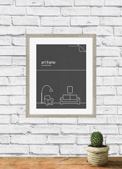 A Deluxe Gallery Frame Ash 40X50CM from Blue Dog Posters, showcasing a minimalist art print with an outline of a living room scene featuring a lamp, chair, and sofa, hangs on a white brick wall. Below the Deluxe Large Frame sits a small potted cactus on a wooden surface. This 40cm x 50cm poster adds elegance to any room.