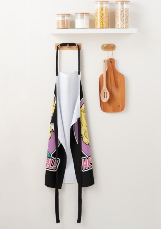 The Ru Paul Hey Kitty Girl Apron by Blue Dog Posters, adorned with vibrant cartoon designs and made from polyester, hangs on a wall hook in the kitchen. Next to the apron, which is part of our homewares collection, a wooden cutting board accompanied by two wooden utensils is also hanging. Above them, a white shelf holds jars filled with various grains and spices.