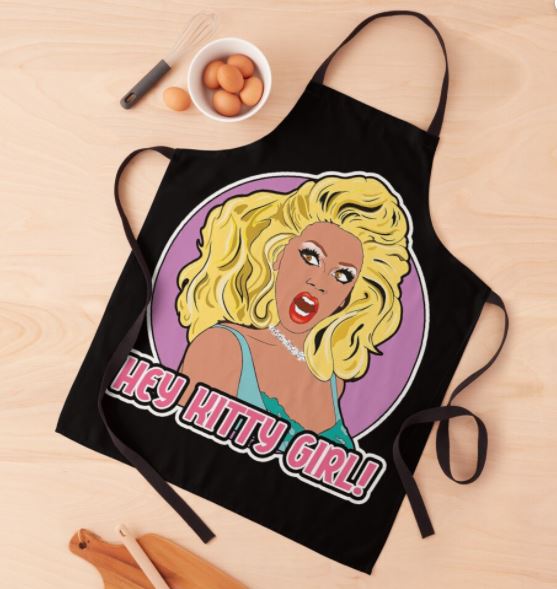 A black Ru Paul Hey Kitty Girl Apron from the Blue Dog Posters Homewares Collection features an illustrated figure of a glamorous person with large blonde hair and a surprised expression. Text below the illustration reads "HEY KITTY GIRL!" The polyester apron is placed on a wooden surface next to a bowl with four eggs and wooden kitchen utensils.