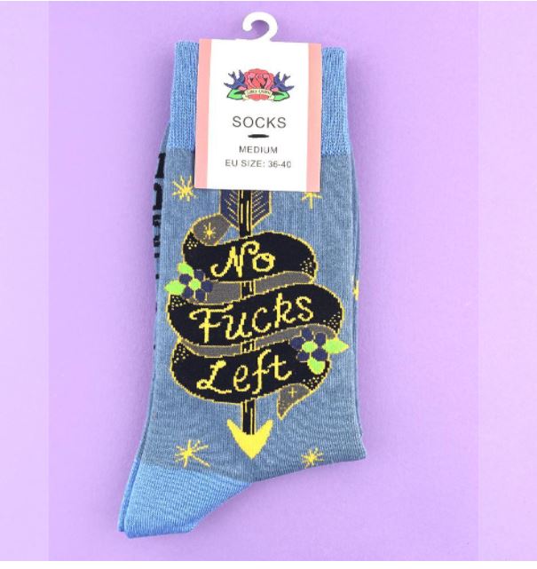 A single grey sock with blue cuffs, featuring a bold yellow and black design of a signpost that reads "No Fucks Left." This Jubly-Umph No Fucks Left Sock by Blue Dog Posters, tagged as medium-sized (EU size 36-40), is unisex and remains attached to its retail tag. The background is a pastel purple.