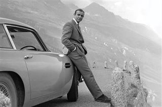 A person in a suit is leaning against a classic car parked on a mountainous road. The backdrop features misty mountains and a winding path. Captured in black and white, the scene evokes a timeless, vintage essence akin to an MTV 045 James Bond - Connery & Aston Martin printed on premium 150gsm gloss paper.