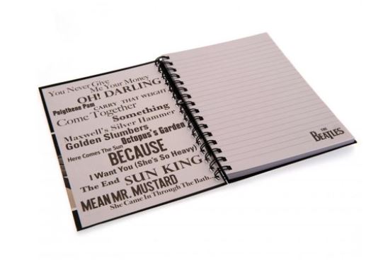 A spiral-bound notebook lies open. The left page features a collage of song titles in various fonts, including "Come Together" and "Oh! Darling." The right page is lined for writing and displays "The Beatles" logo at the bottom corner, making it a perfect addition to any stationery gifts collection. This is the Blue Dog Posters' The Beatles Abbey Road A5 Spiral Notebook.