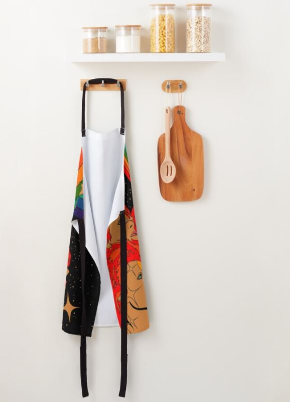 A vibrant Starman by Audrey Herbertson apron from Blue Dog Posters, featuring starry and lion-themed designs, hangs on a wooden hook beneath a simple white shelf. This shelf, part of our Homewares Collection, holds jars of oats and rice. Below it, two wooden cutting boards and a spoon also dangle from hooks.