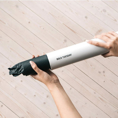 A person holds a cylindrical white tube with the text "Blue Dog Posters" printed on it. The tube has a black cap on one end and is being held over a light wooden background.