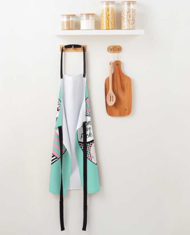 A kitchen wall with a minimalistic design holds a shelf with three jars filled with grains. Below the shelf, a turquoise polyester "I Dont Give a Fuck Apron" from Blue Dog Posters, featuring a colorful pattern, hangs on a hook. Next to the apron, there is a wooden cutting board and a wooden spoon on another hook. The apron is machine washable for easy care.
