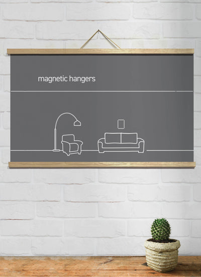 A minimalist drawing of a living room on a dark grey canvas hangs on a white brick wall using Blue Dog Posters Magnetic Timber Hanger 81cm Natural Pair. The artwork showcases a floor lamp, an armchair, and a sofa, with a small potted cactus on a wooden surface below. The text "Magnetic Timber Hanger" is displayed on the canvas.