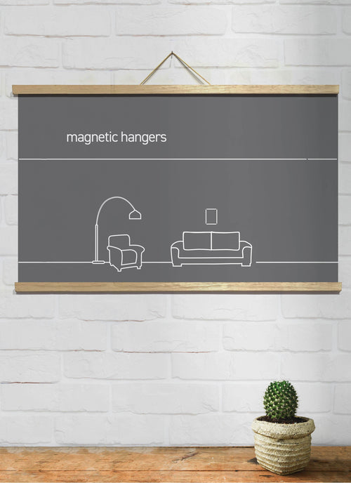 A minimalist drawing of a living room on a dark grey canvas hangs on a white brick wall using Blue Dog Posters Magnetic Timber Hanger 81cm Natural Pair. The artwork showcases a floor lamp, an armchair, and a sofa, with a small potted cactus on a wooden surface below. The text "Magnetic Timber Hanger" is displayed on the canvas.