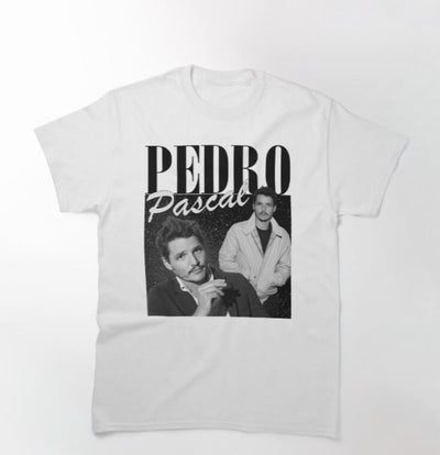 The Pedro Pascal T-shirt White MED from Blue Dog Posters features two black-and-white images of Pedro Pascal, with his name printed in bold, stylized letters above and between the photos. The left image shows him smoking a cigarette, while the right image captures him standing with his hands in his jacket pockets.