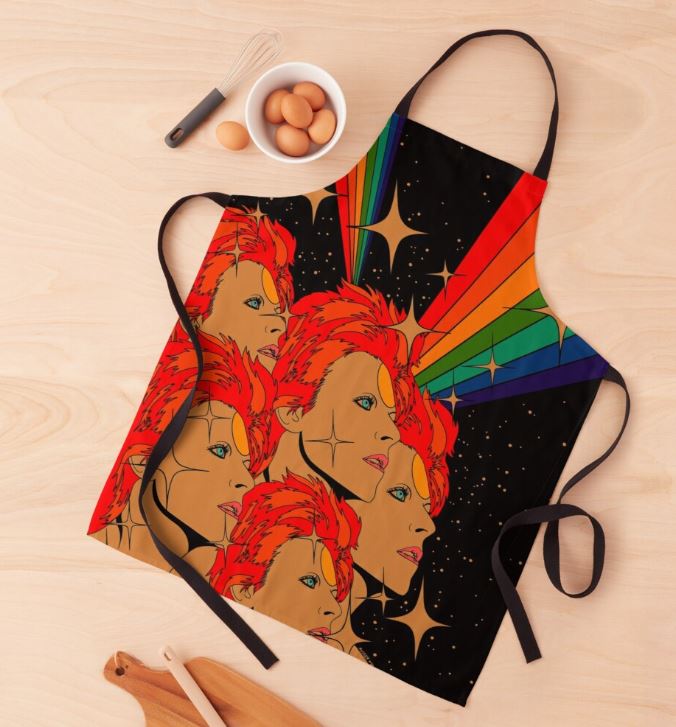 A vibrant Starman by Audrey Herbertson Apron from Blue Dog Posters' Homewares Collection lies flat on a wooden surface. The 100% polyester apron features illustrated faces with red hair against a black background with rainbow beams and starbursts. Nearby, eggs rest in a bowl, accompanied by a whisk and wooden utensils.